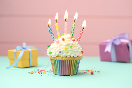 Burning candles in cupcake, sprinkles and gift boxes on turquoise table against pink background