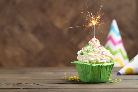 Burning sparkler in cupcake, sprinkles and party hats on wooden table, space for textの写真素材