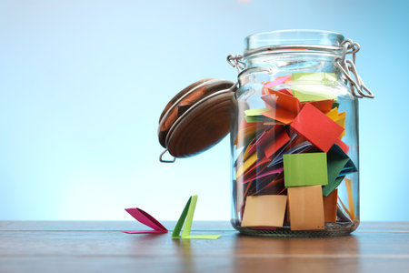 Jar with colorful pieces of paper on wooden table against light blue background. Space for textの写真素材