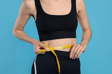 Woman measuring her belly with tape on light blue background, closeup