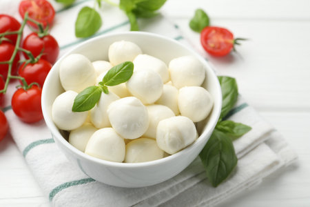 Tasty mozzarella cheese balls in bowl, basil and tomatoes on white wooden table, closeupの写真素材