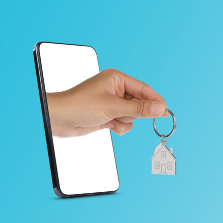 Woman's hand with keychain in shape of house sticking out from smartphone on light blue background. Real estate services onlineの写真素材