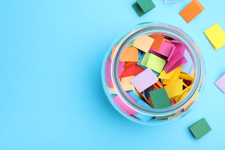 Jar with colorful pieces of paper on light blue background, flat lay. Space for text