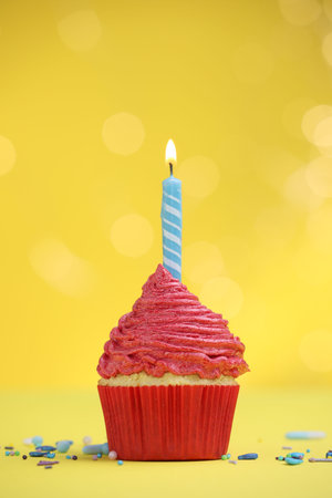 Burning candle in cupcake and sprinkles on yellow background with blurred lightsの写真素材