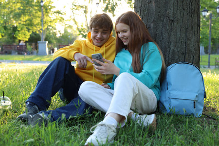 Teenage girl and boy with smartphone on green grass in parkの写真素材