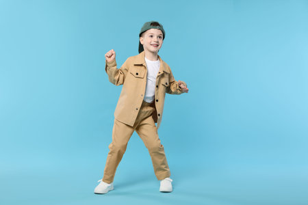 Happy little boy in baseball hat dancing on light blue background