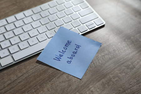Paper note with words Welcome Aboard and computer keyboard on wooden table, closeupの写真素材