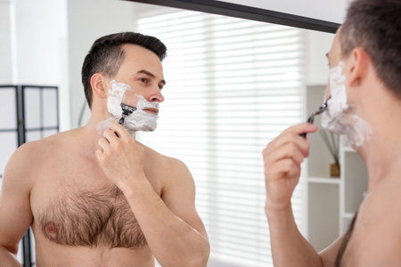 Handsome man shaving with razor near mirror at homeの写真素材