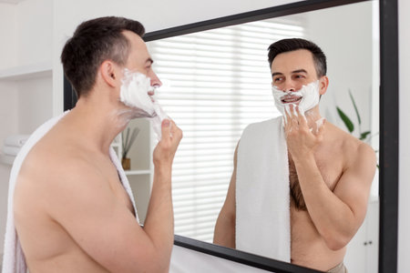 Smiling man applying shaving foam near mirror in bathroomの写真素材