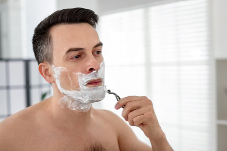 Handsome man shaving with razor near mirror at homeの写真素材
