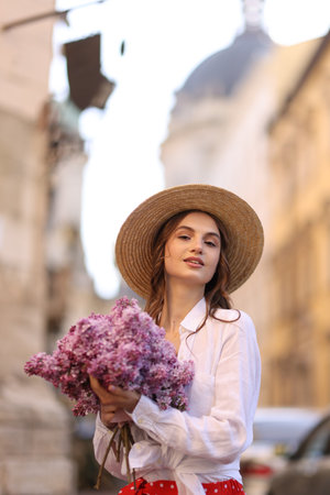 Beautiful woman in hat with bouquet of lilac flowers outdoorsの写真素材