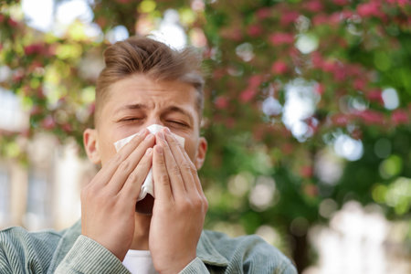 Young man with paper tissue suffering from seasonal pollen allergy near blossoming tree on spring day. Space for textの写真素材