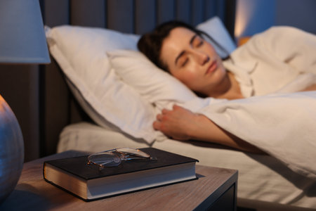Young woman sleeping in bed at home, focus on glasses and book