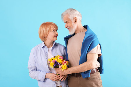 Lovely senior couple with bouquet of flowers on light blue background