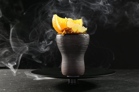 Hookah bowl with citrus fruits and smoke on black table, closeup