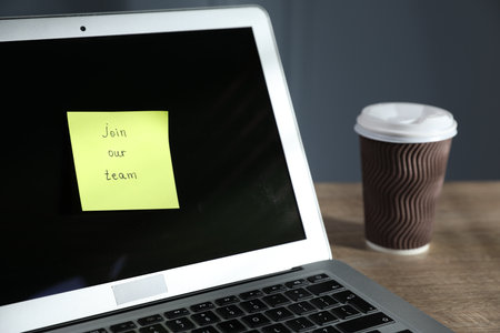 Sticky note with words Join Our Team, laptop and takeaway cup on wooden table against gray background, closeupの写真素材