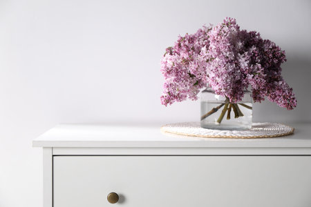 Beautiful lilac flowers in glass vase on cabinet near white wall indoors. Space for textの写真素材