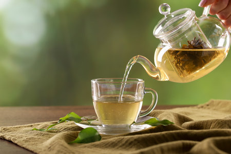 Woman pouring tasty green tea from teapot into cup at table against blurred background, closeupの写真素材