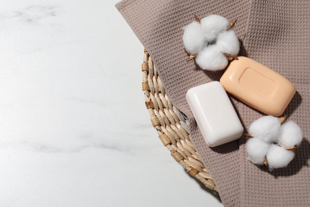 Soap bars, towel and cotton flowers in wicker basket on white marble table, top view. Space for textの写真素材