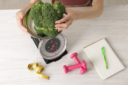 Woman weighing broccoli on kitchen scale at light wooden table indoors, top viewの写真素材