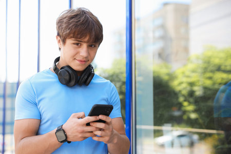 Cute teenage boy with headphones and smartphone near building outdoors