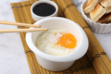 Half-boiled egg in bowl, soy sauce, chopsticks and toasted bread on white marble table, closeup. Traditional asian breakfastの写真素材