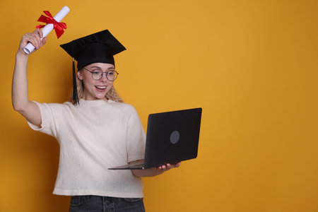 Happy student with diploma and laptop after graduation on orange background. Space for textの写真素材