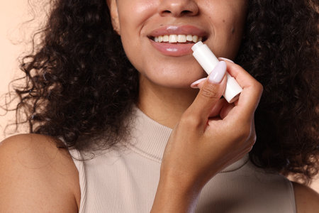 Smiling woman applying lip balm onto her lips on beige background, closeup