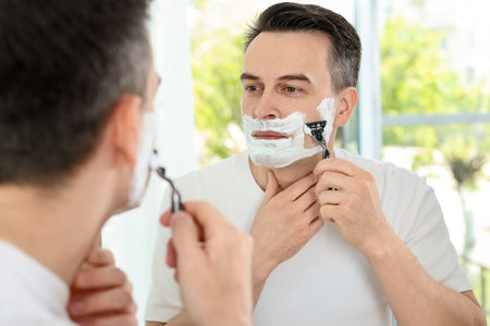 Handsome man shaving with razor near mirror at homeの写真素材