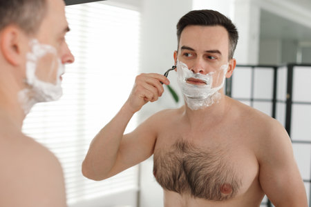 Handsome man shaving with razor near mirror at homeの写真素材