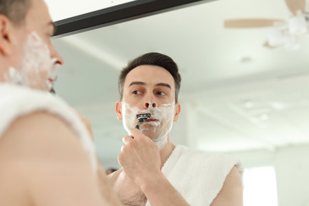 Handsome man shaving with razor near mirror at home, low angle viewの写真素材