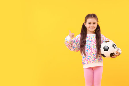 Little girl with soccer ball showing thumbs up on yellow background, space for textの写真素材