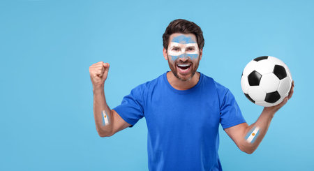 Excited fan with flags of Argentina painted on his arms holding soccer ball against light blue background. Banner design with space for textの写真素材
