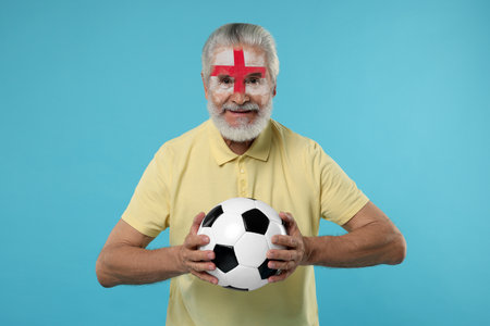 Fan with flag of England painted on his face holding soccer ball against light blue backgroundの写真素材