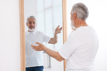 Handsome senior man looking at himself in mirror indoorsの写真素材
