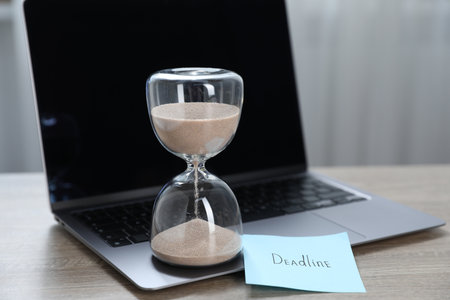 Sticky note with word Deadline, hourglass and laptop on wooden table indoors, closeupの写真素材