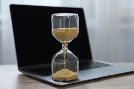 Deadline. Hourglass with flowing sand and laptop on wooden table indoors, closeupの写真素材