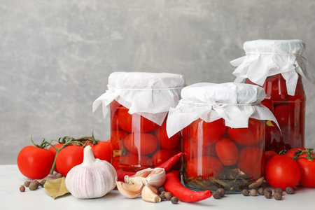 Tasty pickled tomatoes in jars, fresh products and spices on white table against gray background, closeupの写真素材