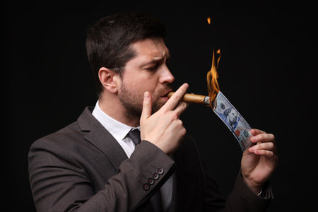Greedy businessman lighting cigar with dollar banknote on black backgroundの写真素材