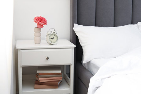 Bedside table with alarm clock and flower near bed indoors