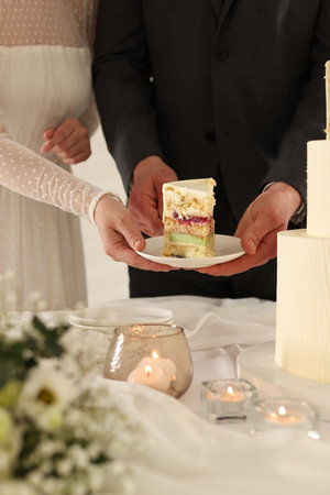 Newlyweds putting plate with piece of wedding cake onto table indoors, closeupの写真素材