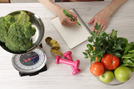 Woman developing diet plan at light wooden table with kitchen scale, measuring tape, dumbbells and healthy food, top viewの写真素材