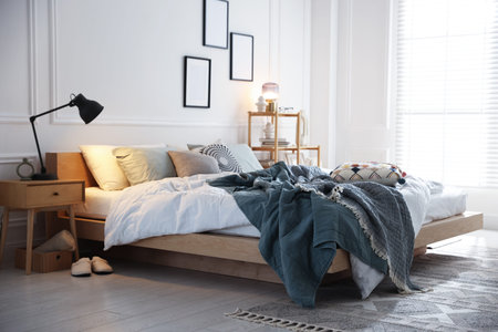 Cozy bed with soft lines, nightstand and lamp in stylish room. Home textile