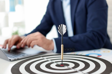 Man working on laptop at table, focus on dartboard with arrow indoors