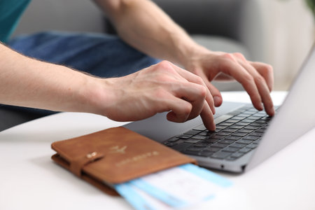 Man using laptop at table with passport and flight tickets indoors, closeup