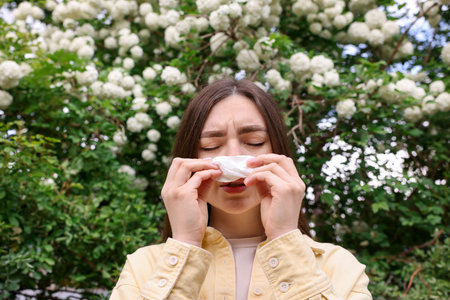 Woman with paper tissue suffering from seasonal pollen allergy near blossoming shrub on spring dayの写真素材