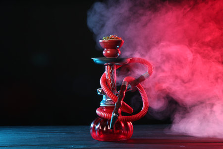 Hookah on wooden table with smoke in color lights against black background