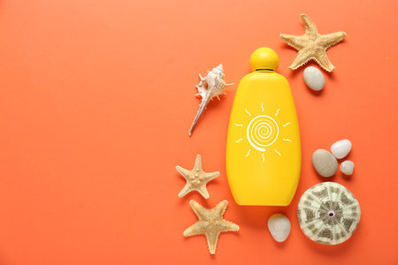 Sunscreen product, shells, sea stars and pebbles on orange background, top view. Bottle with drawing of sunの写真素材