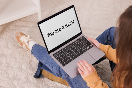 Cyber bullying. Upset teenage girl and laptop with text You Loser on floor indoors, closeupの写真素材