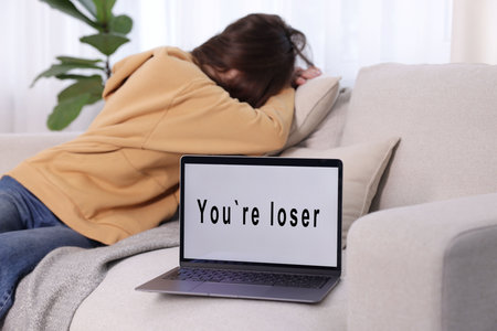 Cyber bullying. Upset teenage girl and laptop with text You Loser on sofa at home, selective focusの写真素材
